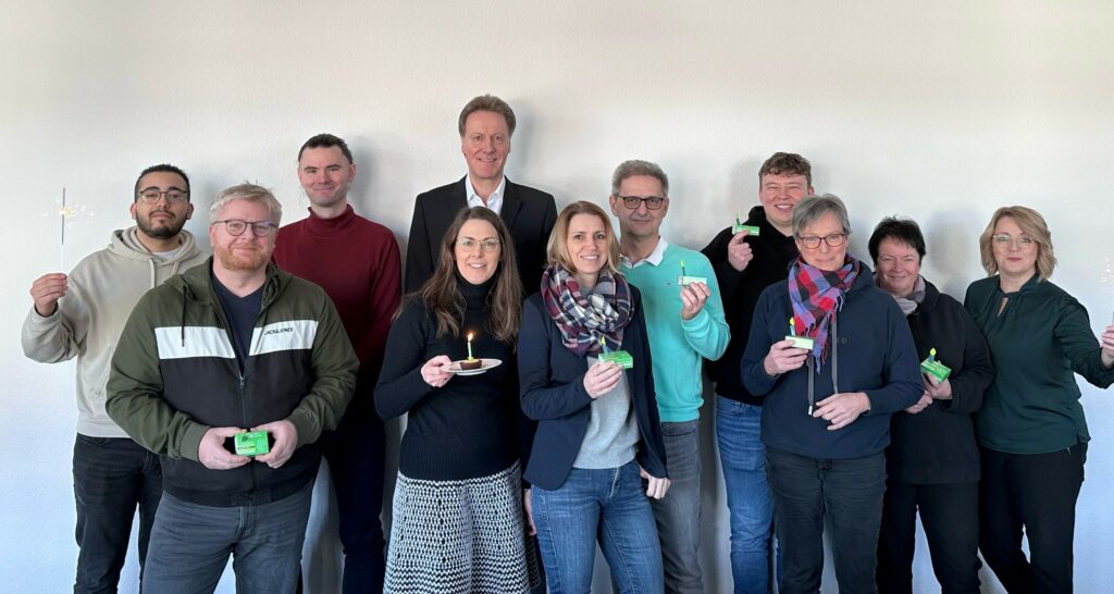 Eine Gruppe von elf Erwachsenen steht in einem Innenraum vor einer schlichten weißen Wand und lächelt in die Kamera. Zur Demonstration der Kooperation halten mehrere Personen kleine grüne Objekte oder Modelle in der Hand, während zwei Personen windmühlenähnliche Gegenstände auf Stöcken präsentieren. - NG.network