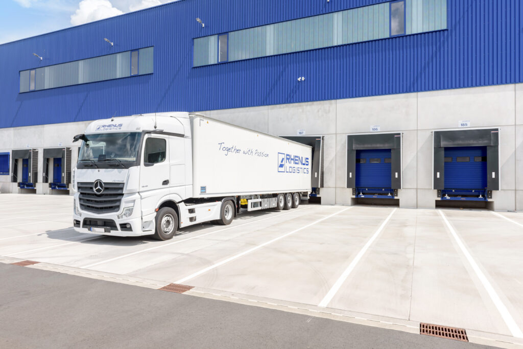 Ein weißer Mercedes-Benz-Sattelzug mit einem Auflieger mit der Aufschrift "Rhenus Logistics" steht an einer Laderampe vor einer großen blauen Industrielagerhalle, die auf Präzision und Kooperation im Stückgutverkehr hinweist. Im Hintergrund sind vier Verladetore zu sehen. - NG.network