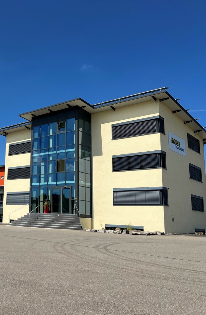 Ein modernes dreistöckiges Bürogebäude mit großen Glasfenstern, hellgelben Außenwänden und einem sichtbaren "STIEGER"-Schild an der Fassade fällt als Drehscheibe der Logistik-Kooperation auf. Eine Betontreppe führt zum Haupteingang. - NG.network