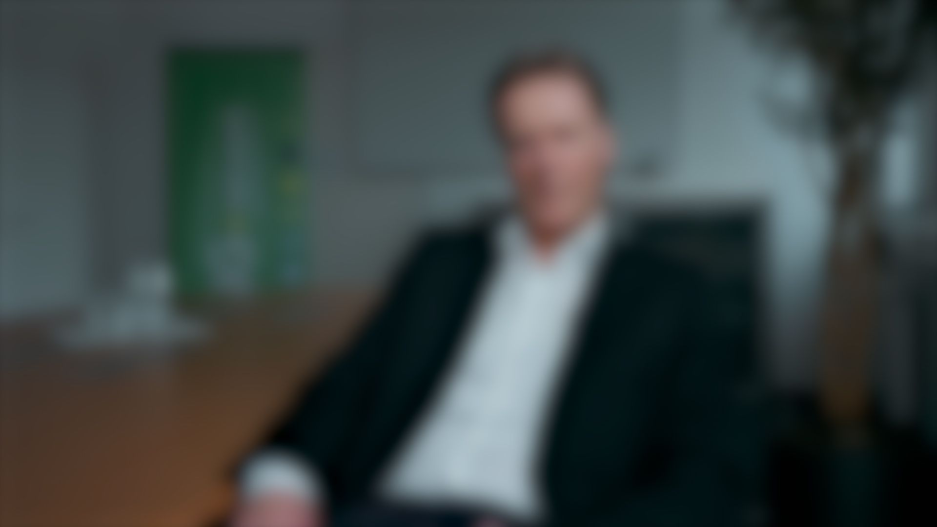 Ein Mann im Anzug sitzt an einem Tisch in einer modernen Büroumgebung. Das Bild ist absichtlich unscharf, wobei die Präzision in der Komposition trotz undeutlicher Details offensichtlich ist. - NG.network