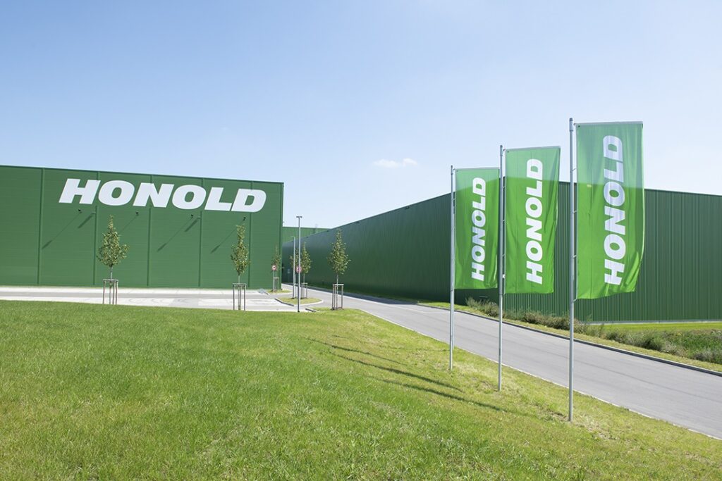 Ein grünes Industriegebäude mit dem Schriftzug "HONOLD" in großen weißen Buchstaben, drei grüne Fahnen mit der Aufschrift "HONOLD", eine gepflasterte Straße und eine Rasenfläche unter strahlend blauem Himmel - das spiegelt Präzision in der Transportlogistik wider. - NG.network