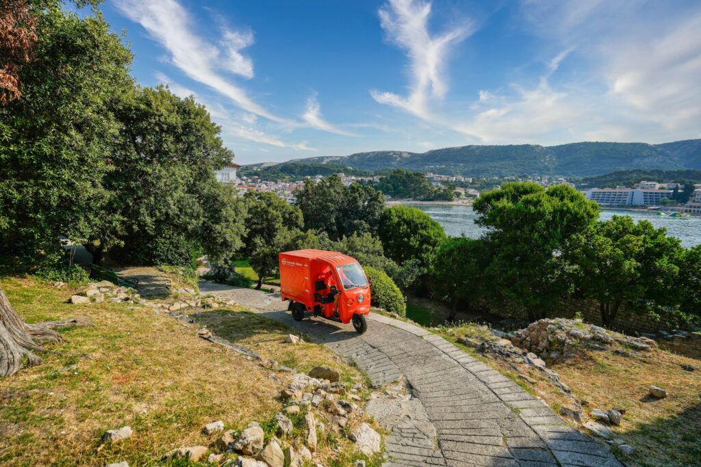 Ein kleiner roter Lieferwagen fährt auf einem steinernen Weg im Grünen in Deutschland, mit Hügeln, Gebäuden und einem Gewässer im Hintergrund unter einem blauen Himmel mit Wolkenfetzen. - NG.network