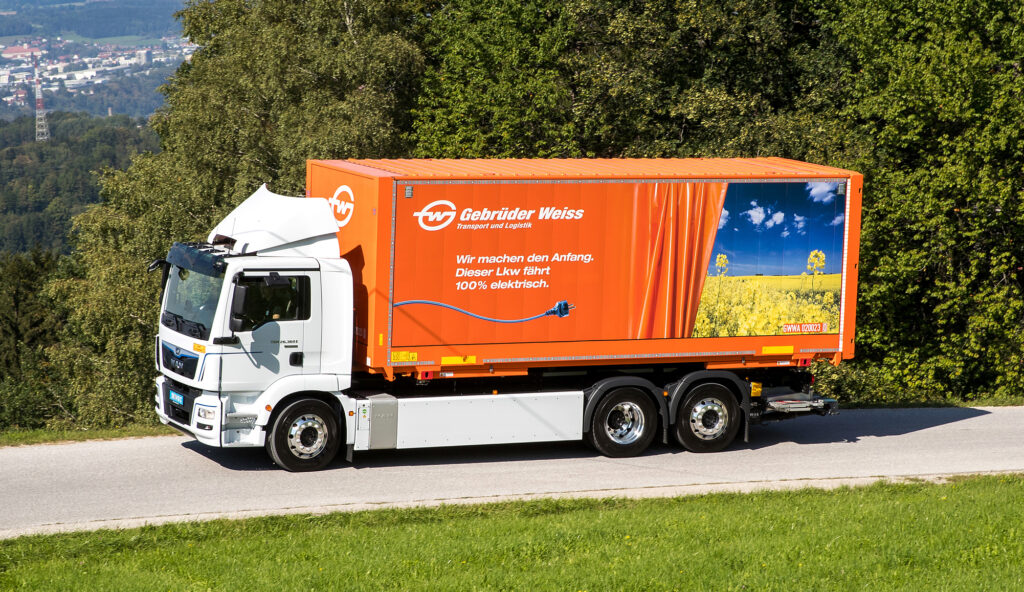 Ein großer weißer und oranger Elektro-LKW mit dem Gebrüder Weiss-Logo und dem deutschen Text über Stückgut auf der Seite fährt auf einer Straße neben grünen Bäumen und Gras. Im Hintergrund sind eine Stadt in Deutschland und entfernte Hügel zu sehen. - NG.network