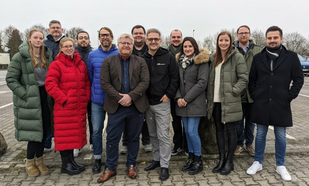 Eine Gruppe von dreizehn Erwachsenen steht in Winterkleidung im Freien auf einem gepflasterten Platz in Deutschland. Sie stehen lächelnd in zwei Reihen; im Hintergrund sind blattlose Bäume und geparkte Autos zu sehen, die Präzision verkörpern. - NG.network
