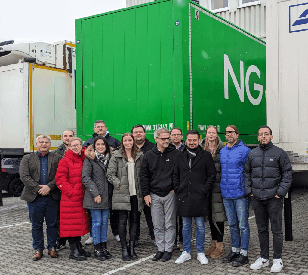 Eine Gruppe von 14 Personen, einige in Wintermänteln, steht im Freien vor einem großen grünen Industriecontainer mit der Aufschrift "NG". Im Hintergrund sind weitere Lastwagen und Container der Firmen Transport und Stückgut zu sehen. - NG.network