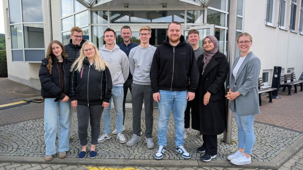 Zehn Personen stehen in zwei Reihen vor einem Gebäudeeingang und verkörpern die Kooperation. Einige lächeln, und die meisten tragen legere Kleidung mit Jacken oder Pullovern. Eine Frau trägt ein Kopftuch, und die Gruppe steht auf gemustertem Pflaster. - NG.network