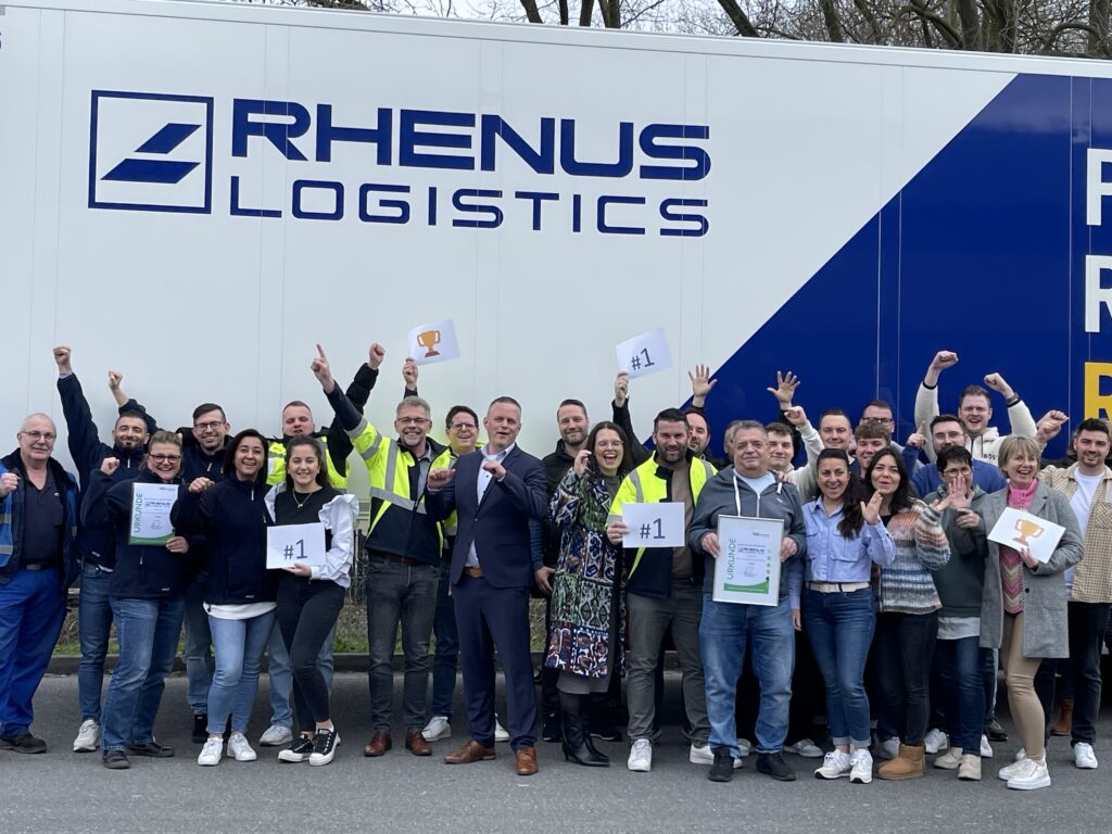 Eine Gruppe von Menschen posiert und lächelt vor einem Lkw mit der Aufschrift "Rhenus Logistik". Einige halten Schilder mit "#1" und Trophäen und einige tragen reflektierende Sicherheitswesten, um ihre Kooperation in Transport und Logistik zu feiern. - NG.network