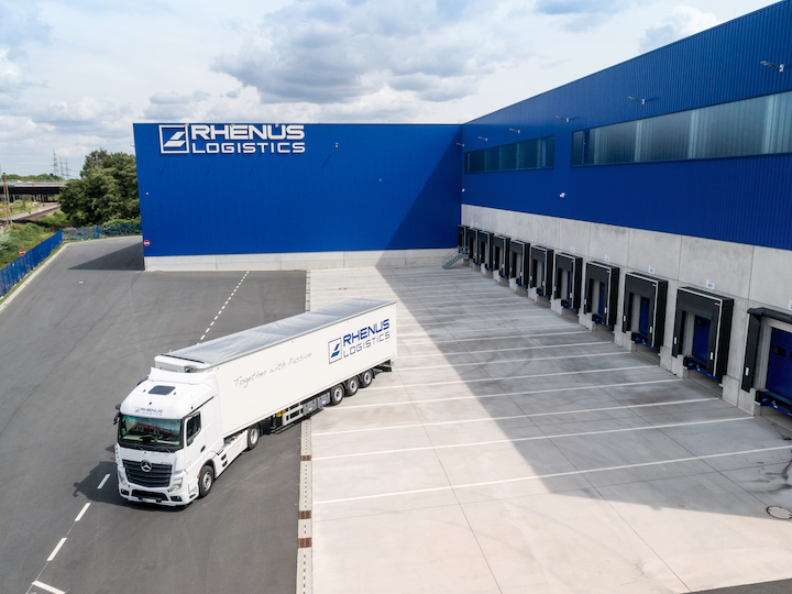 Ein weißer Lkw von Rhenus Logistics steht vor einem großen blauen Lagergebäude mit mehreren Ladebuchten und präsentiert Präzision in Transport auf einem größtenteils leeren Asphaltplatz unter einem teilweise bewölkten Himmel in Deutschland. - NG.network