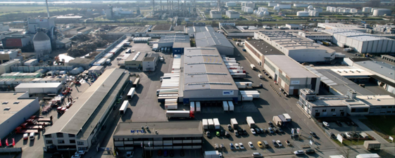 Luftaufnahme eines Industriegebiets in Deutschland mit mehreren Lagerhäusern und Fabriken. Zwischen den Gebäuden verlaufen Straßen, auf denen Lastwagen, Autos und Lagertanks geparkt sind - ein Beispiel für Präzision und Kooperation in der Logistik. - NG.network