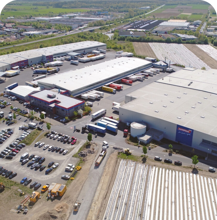Luftaufnahme eines Industriekomplexes mit großen Lagerhallen, Stückgutbetrieben, geparkten Lastwagen an Laderampen, Baufahrzeugen und angrenzenden landwirtschaftlichen Feldern. Straßen und Grünflächen umgeben das Gelände und unterstreichen die Präzision bei der Gestaltung und Logistik. - NG.network