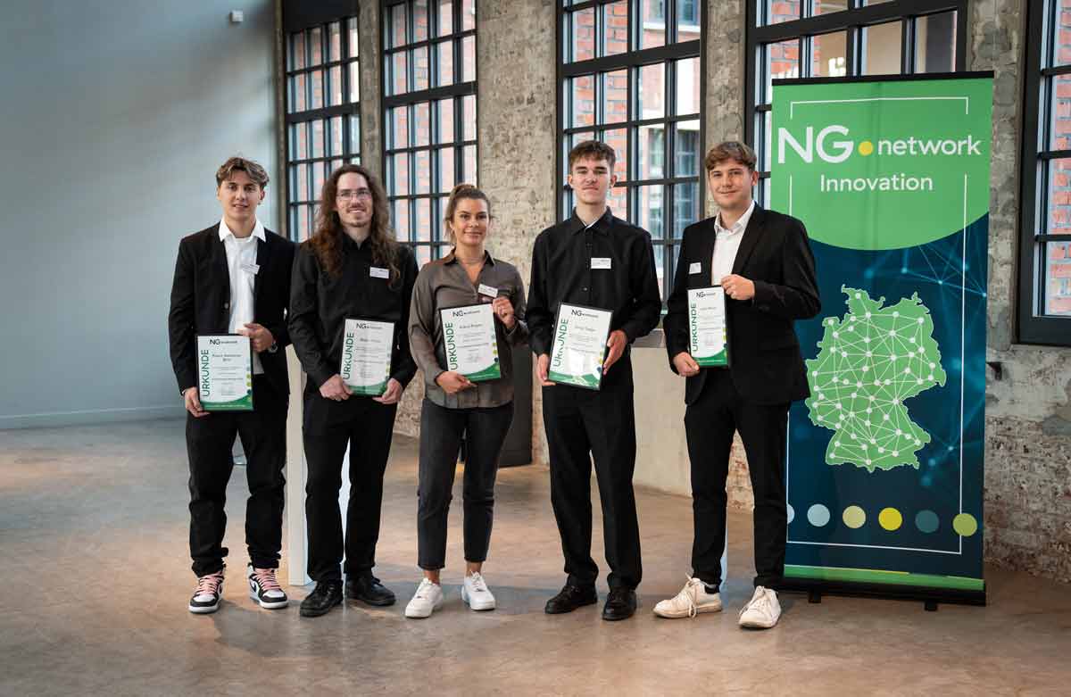 Fünf Personen stehen drinnen vor einem großen Banner von NG Network Innovation und einer Backsteinmauer mit Fenstern, jede hält ein Zertifikat und trägt ein Namensschild. Vier von ihnen tragen Geschäftskleidung, einer ein graues Hemd und weiße Turnschuhe, die Präzision im Stückgut demonstrieren. - NG.network