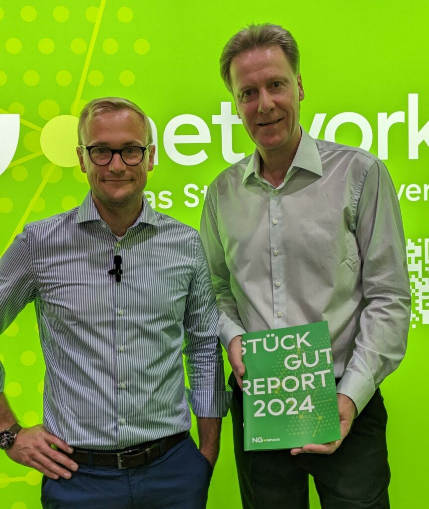 Zwei Männer stehen vor einem hellgrünen Hintergrund mit weißer Schrift. Einer hält ein Buch mit dem Titel "STÜCK GUT REPORT 2024" in der Hand. Beide, gekleidet in Button-up-Hemden, repräsentieren Stückgut Präzision in Deutschland und schauen direkt in die Kamera. - NG.network