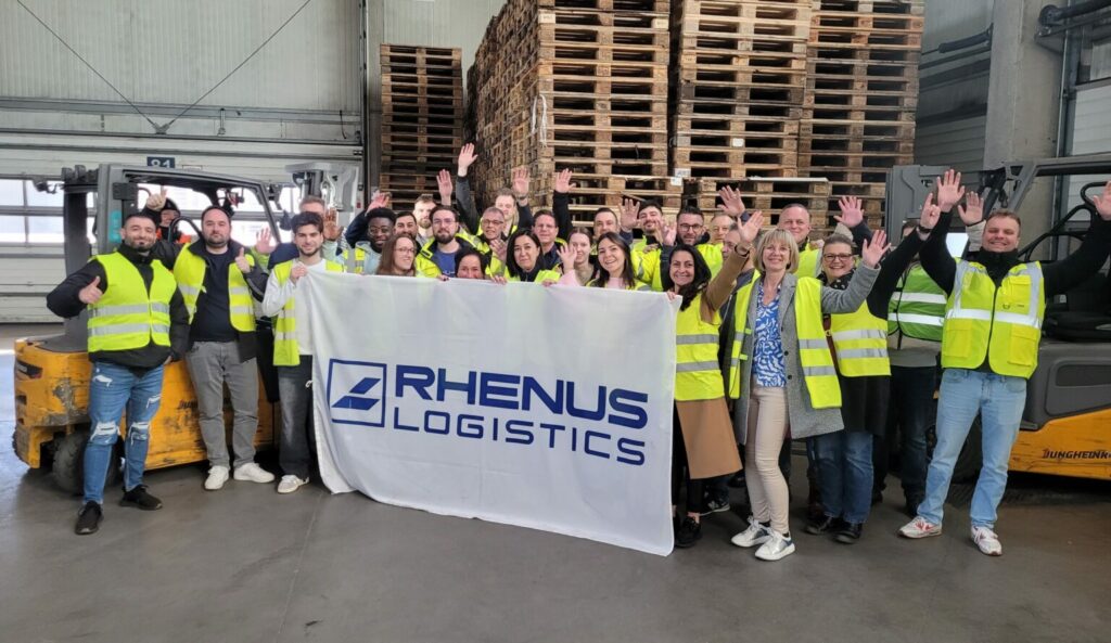 Eine Gruppe von Menschen in gelben Sicherheitswesten steht in einer Halle in Deutschland und hält lächelnd und winkend ein großes "Rhenus Logistics"-Transparent vor gestapelten Holzpaletten und Industrieanlagen, die auf Präzision im Transportwesen hinweisen. - NG.network
