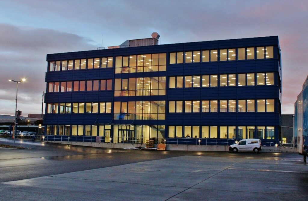 Ein vierstöckiges modernes Bürogebäude mit großen beleuchteten Fenstern in der Abenddämmerung stellt sein Engagement für Nachhaltigkeit unter Beweis. Ein Lieferwagen ist in der Nähe des Eingangs geparkt, und auf dem nassen Boden spiegeln sich die Lichter des Gebäudes vor einem wolkenverhangenen, sonnenuntergangsfarbenen Himmel. - NG.network