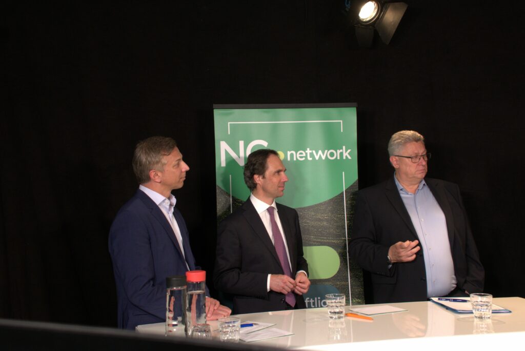 Drei Männer in Business-Kleidung stehen an einem weißen Tisch mit Wassergläsern und einer roten Flasche. Hinter ihnen hebt ein grünes "NG network"-Schild ihre Kooperation hervor, während der dunkle Hintergrund und das Scheinwerferlicht die Präzision der Szene verstärken. - NG.network