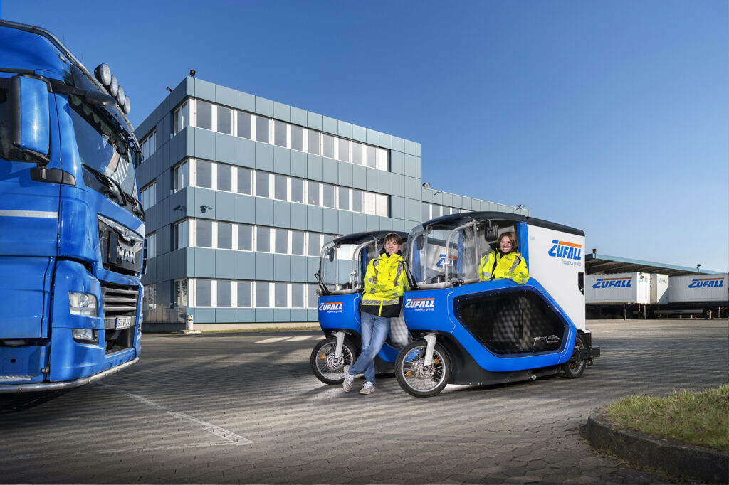 Zwei Personen in gelben Sicherheitswesten stehen neben blau-weißen Elektro-Lieferfahrzeugen mit der Aufschrift "ZUFALL", die auf Nachhaltigkeit hinweisen, vor einem modernen Industriegebäude und einem blauen Lkw auf einem gepflasterten Parkplatz. - NG.network
