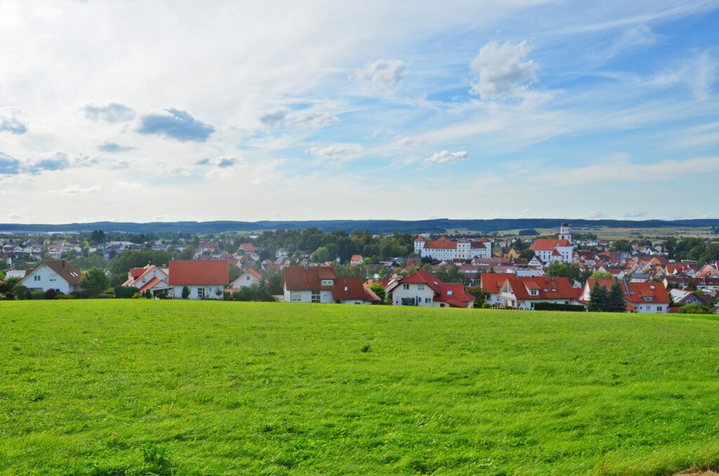 Ein grasbewachsenes Feld im Vordergrund mit einer kleinen Stadt aus weißen Häusern mit roten Dächern im Mittelgrund, die Präzision in ihrer Anordnung zeigt, umgeben von Bäumen unter einem teilweise bewölkten Himmel. - NG.network