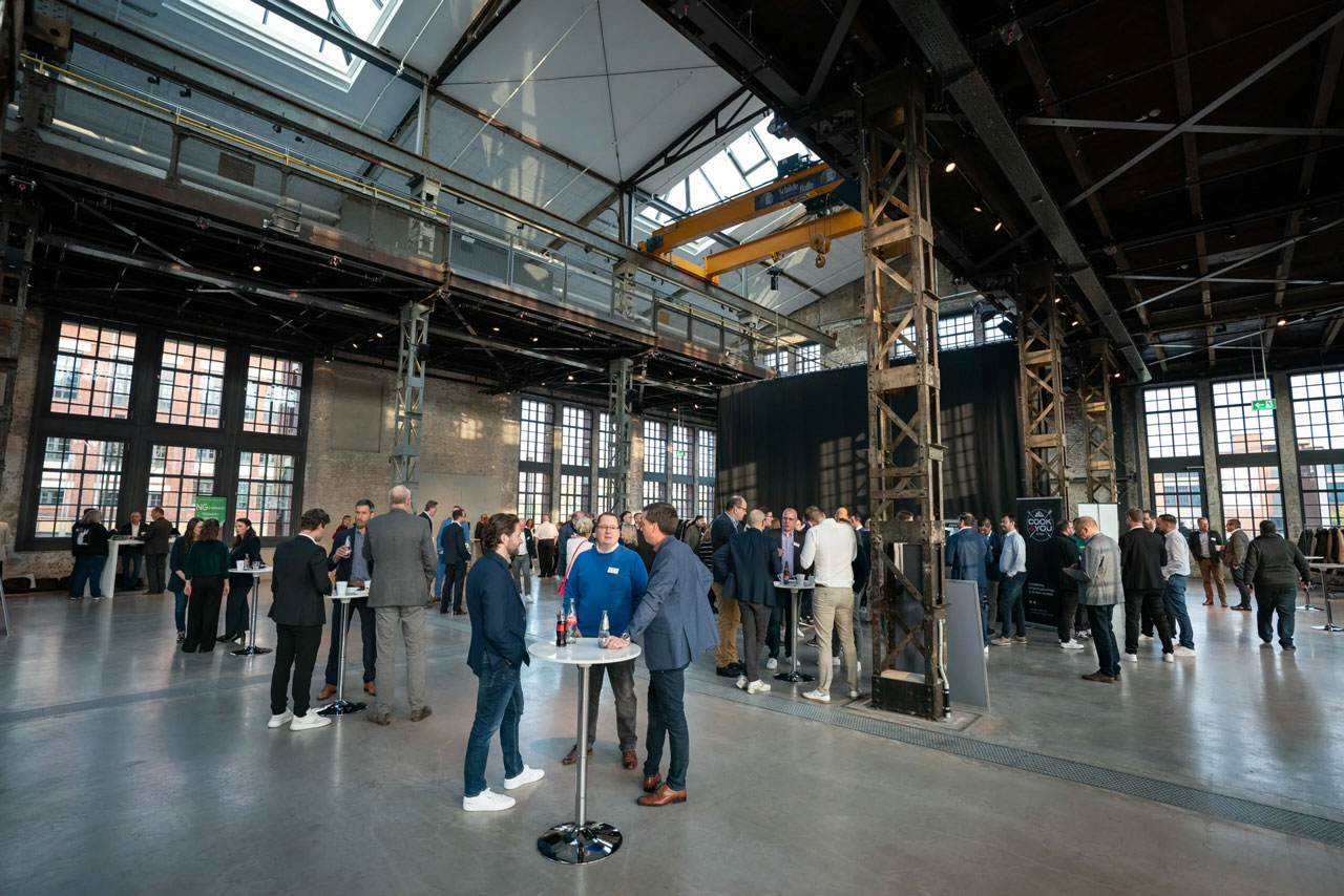 In einem großen Veranstaltungsraum im Industriestil in Deutschland mit hohen Decken, freiliegenden Balken und großen Fenstern stehen mehrere Personen in kleinen Gruppen und unterhalten sich. Einige stehen mit Getränken um hohe Tische herum. - NG.network